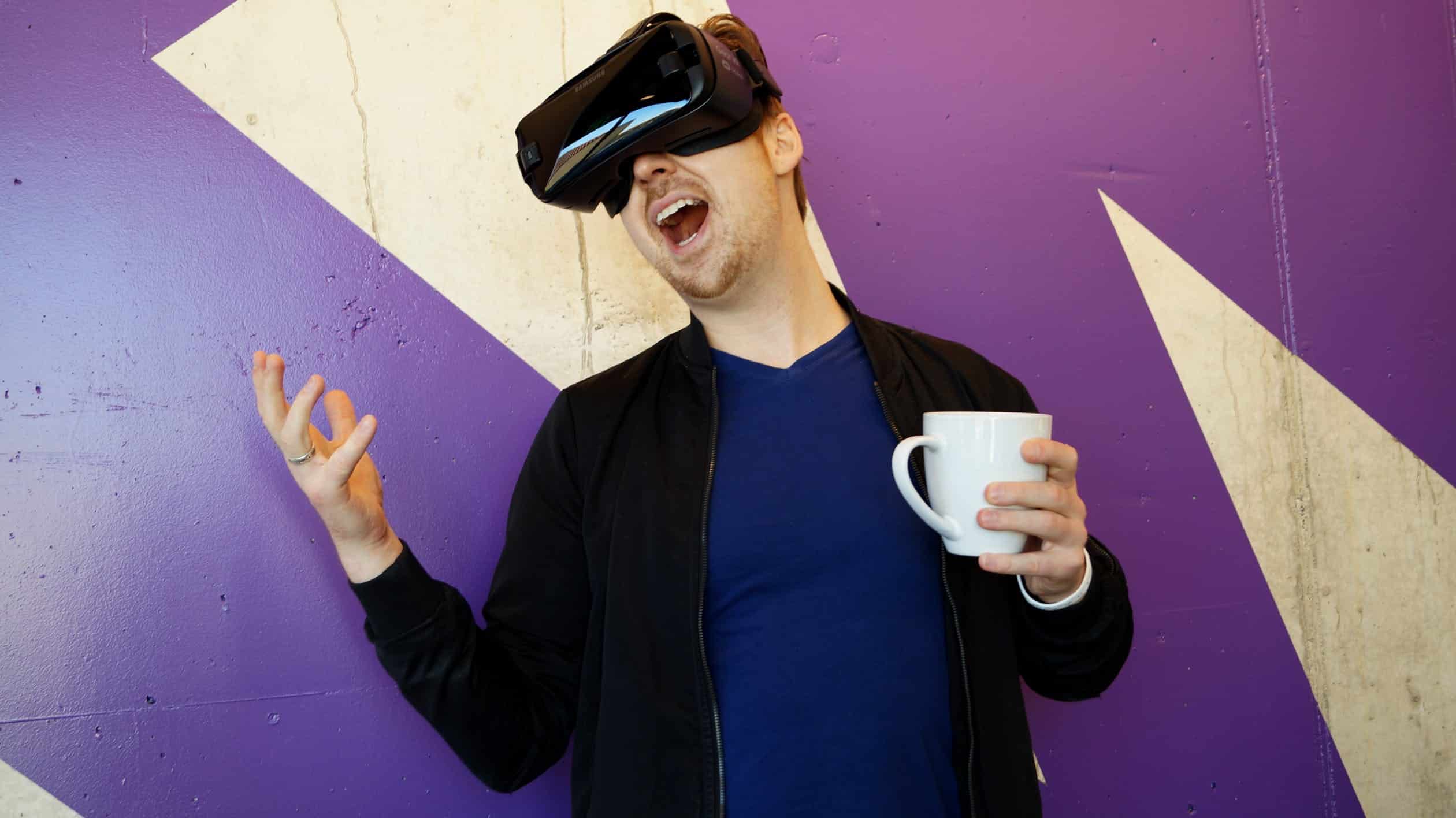 A man wearing virtual reality headset and holding a mug, expressing excitement in front of a purple wall