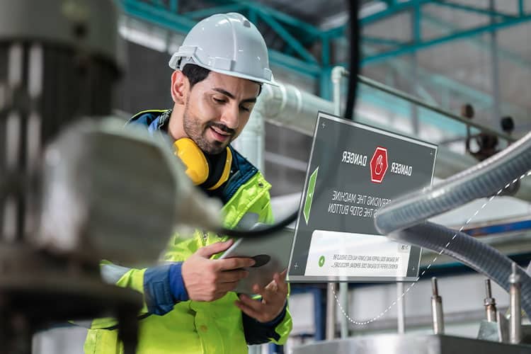 A construction worker wearing a hard hat, safety vest, and protective gear uses a digital tablet on a construction site.