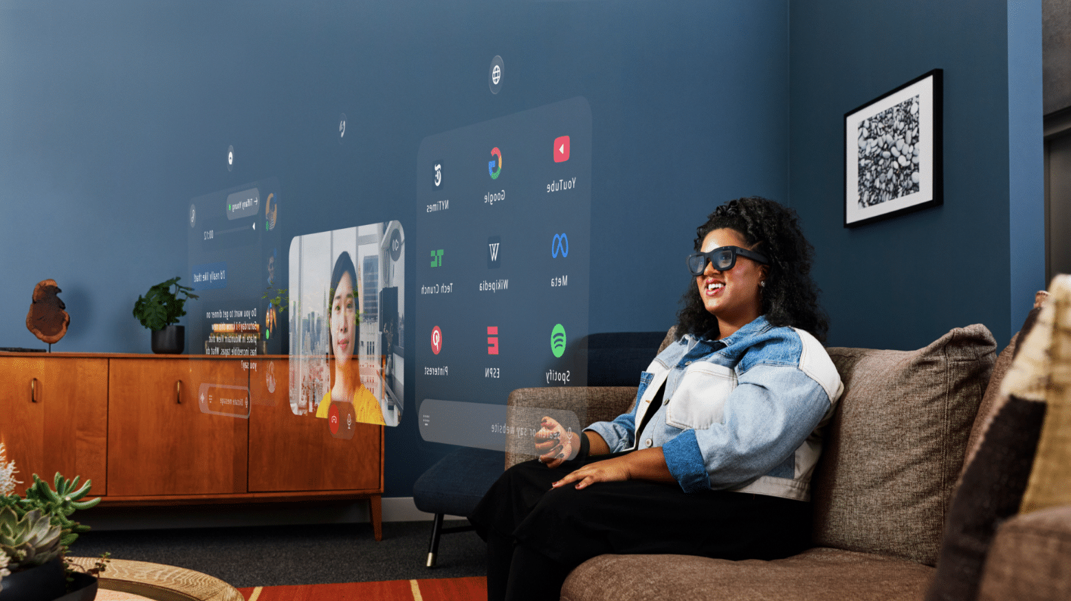 Woman wearing glasses and a denim jacket sitting on a couch in a room with technology-themed wall art