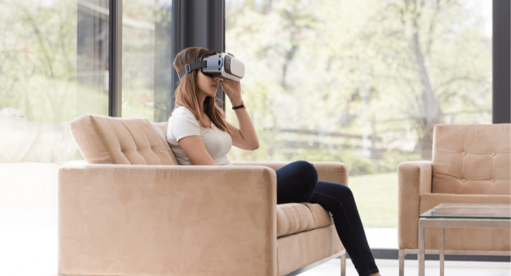 Woman wearing virtual reality headset sitting on couch in front of large window with nature view
