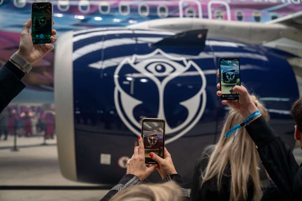 Two people photographing with their smartphones in front of a large video screen displaying an app interface and the logo of an event or organization.