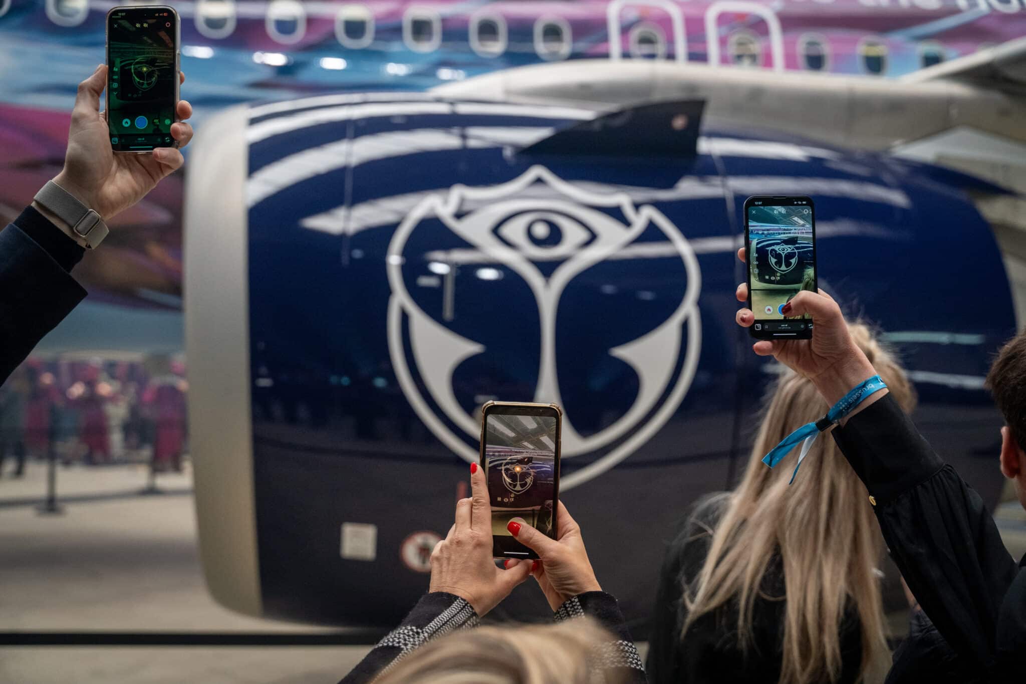 Two people photographing with their smartphones in front of a large video screen displaying an app interface and the logo of an event or organization.